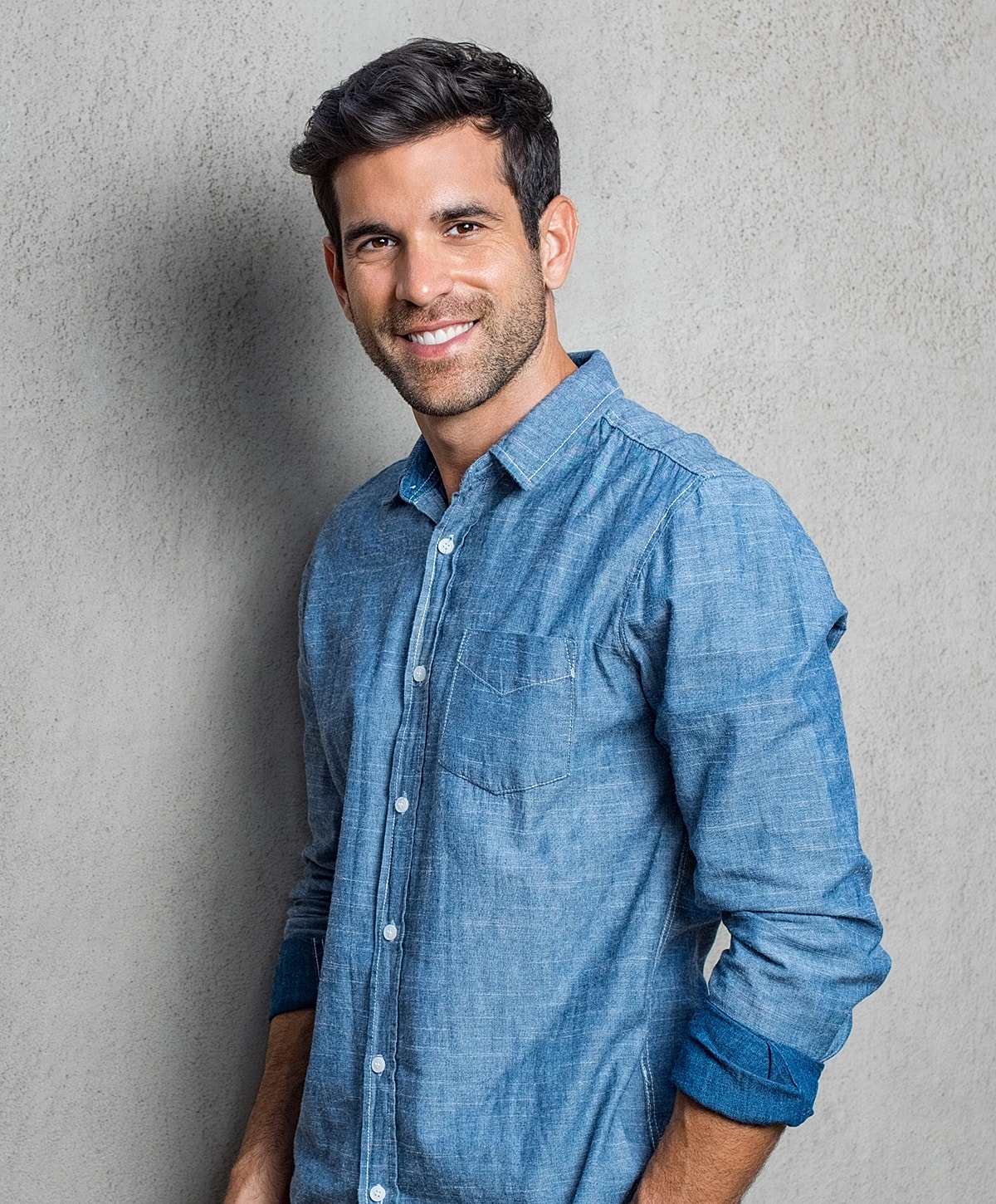 Smiling man in a denim shirt against gray wall.